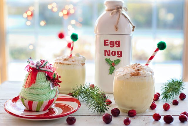 Image of festive table containing a bottle of eggnog, 2 glasses of eggnog decorated with whipped cream, and a cupcake